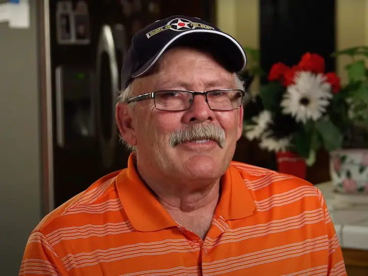 Senior man with mustache smiling wearing a baseball cap and orange striped polo shirt